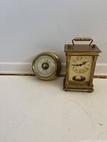 Barometer and small brass carriage clock side view on a white surface.
