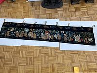 Full front view of the black embroidered tapestry cloth laid out flat, showing detailed colorful embroidery of traditional figures, animals and plants with hanging loops.