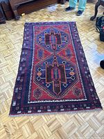 Full view of rectangular Afghan rug lying flat on parquet floor showing geometric medallion designs in blue, red, and dark colors with patterned border.