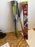Photo showing two large boxed kites standing upright with visible packaging and graphics against white wall on parquet floor