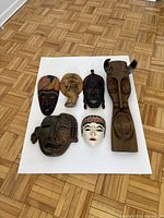 Seven masks arranged on floor with six wooden and one white ceramic mask