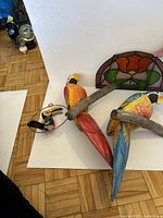 Three wooden parrots perched on branches with a stained glass panel in the background, showing the full parrot figures and part of the stained glass panel.
