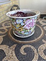 Photo showing side view of round ceramic plant pot with colorful flowers and golden dragon design, containing dried plant material, on mosaic table.