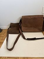 Two brown leather satchels placed on white surface against a wall: medium satchel with flap and adjustable strap in front, smaller satchel behind.