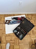 Photo of Stanley manual hand saw, reciprocating jig saw, and an open black case with drill bits and small tools on parquet floor