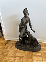 Full side view of the faux bronze sculpture showing the woman kneeling with dog on an oval base against a white wall with parquet flooring.