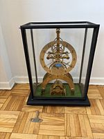 Front view of brass skeleton clock in black metal and glass case on green felt base with winding key placed in front