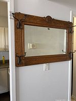 Full view of wall-mounted oak wood frame featuring a rectangular mirror and four black metal double coat hooks, two on each vertical side near corners.