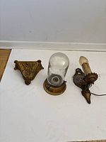 Three vintage wall lights displayed on the floor: ornate brass/bronze sconce, dome glass wall light, candle-style wall light.