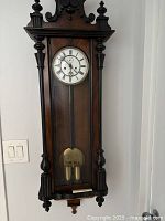 Frontal view of Vienna regulator wall clock showing white clock face with Roman numerals, pendulum, and brass weights behind glass case.