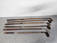 View of six antique wooden shaft golf clubs laid flat showing metal club heads, lengths, and varying grips.