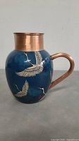 Front and side view of the copper jug showing the blue body with crane bird artwork and copper handle.