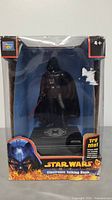 Photo showing the Star Wars Darth Vader electronic talking bank in its original packaging. Bank is a black plastic Darth Vader figure on a base with Galactic Empire insignia. Box has colorful Star Wars themed graphics and a 'Try Me!' feature.