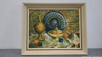 Framed oil painting showing still life with a brass pitcher, blue patterned plate, and various fruit on a white cloth with a textured wall background.