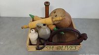Photo of a wooden crate labeled 'Fresh Olives' containing two wooden rolling pins, three jugs of ceramic or stoneware, and a cast iron kitchen tool with handle.