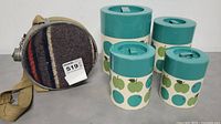 Vintage fabric-covered canteen with beige strap next to four Mid-Century Modern style canisters featuring green and teal apple designs on cream background.