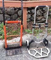 Both hand trucks side by side on concrete near a wooden structure and stone wall, showing orange metal and silver aluminum frames with wheels and base plates.