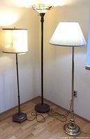 Full view of all three floor lamps. Middle lamp has stained glass shade with pink flowers. Brass lamp on right has white pleated shade. Vintage style brass plated lamp on left has square base and white fabric shade.