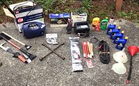 Wide angle photo showing all items in the lot laid out on ground, including battery charger, fluids, hand tools, jumper cables, funnels, and the electric pressure washer box.