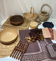 Overview of lot showing woven placemats, multiple woven baskets, beige and dark wood bowls, wooden napkin rings, round woven containers with lids, a folded patterned tablecloth, and napkins.