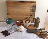 Full lot view including butcher block, meat cleaver, mortar and pestle, garlic keeper, beer stein, fondue set with forks, iron, and water dipper on table.