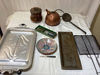 Lot overview showing kettle, jar, trays, skillet, utensils, enameled dish, grilling basket, and skimmer on white cloth