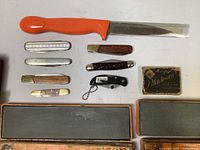 Full view of collection showing Gerber fillet knife, multiple pocket knives of varying handle material, Blakiewicz blade box, and two sharpening stones.