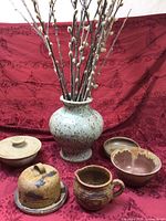 Full view of pottery set on red fabric background showing all items including the tall vase with pussy willow branches and various kitchenware pieces arranged around it.