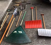 Photo showing various long-handled yard tools including rakes, shovels, a snow shovel, and scrapers laid on pavement.
