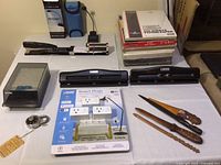 Overall view of table showing paper cutter, stapler, black paper punchers, letter openers, new smart plugs, envelopes, and packs of typing paper.