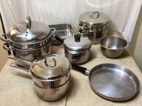 Full view showing stainless steel multi-tier steamer pot, various saucepans with lids, frying pan, perforated insert, and metal mixing bowl.