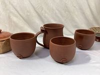 Front-left view showing four Daily Bird earthenware cups in reddish brown, including one with a handle and one handless cup with a crack; Redwing lidded bowl and Nicodemi bowl in background.