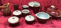 Overview of all pottery pieces including casseroles, mugs, covered pots, and pitchers on red patterned cloth.