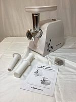 White Sunmile SM-G35 meat grinder unit with attached metal grinding head and two white plastic sausage tubes beside it. Instruction manual and grinding plate accessory sealed in plastic also visible.
