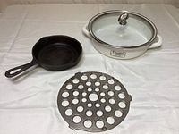 White enameled saut pan with glass lid, black iron skillet, and cast iron trivet displayed on table