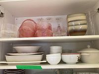 Shelf containing pink and clear textured glass bowls, clear stemware glasses, and striped beige pottery bowls stacked on the right side, with white and decorated bowls below.