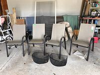 Four outdoor metal frame chairs with beige weather resistant fabric seats and backs arranged in a garage with two black metal mesh baskets placed in front of them.