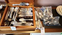Flatware set displayed in wooden box with pieces wrapped in protective plastic and additional flatware sealed in bags placed on adjacent surface.