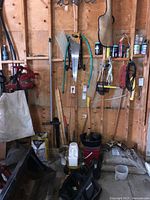Wide shot of garage wall showing large saw with wooden handle, garden hoses, various garden tools, and bags of soil on floor.