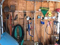 Multiple long-handled garden tools hanging on garage wall including shovels, rakes, hoes, pitchfork, hose coils, bow saw, loppers, hedge shears, pruning shears, face shield.