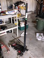 Motomaster garage floor jack positioned vertically in a garage setting with shelving and various items in background