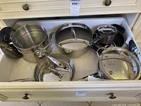 Drawer open showing stainless steel pots, pans, skillets, and lids with reflective surfaces and some wear.