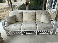 Full front view of white rattan sofa with five cushions and two throw pillows on a carpeted porch area with a backyard view.