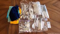Spread of mostly silverplated flatware arranged on a wooden table including forks, knives, spoons, and salad tongs mostly in plastic bags alongside small fabric bags.
