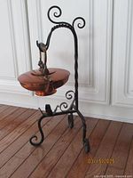 Copper pot hanging on black wrought iron stand with scroll curlicue design on feet, photographed on wooden floor against paneled white wall.