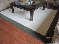 Top view of a beige and green border carpet under a glass-top coffee table on a hardwood floor, showing the carpet's size and condition.