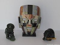 Front view of three items: the large striped semi-precious stone mask in center on black base, nephrite jade kneeling man figurine holding a mirror plate on left, and jade grizzly bear figurine on right.
