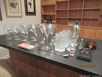 Assorted martini, cognac glasses, shot glasses, frosted mugs, and two ceramic mugs displayed on counter with bar shelves in background.
