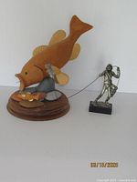 Wide shot showing the largemouth bass wooden sculpture with smaller fish, mounted on a wooden base, next to the pewter fly fishing figurine on a black base.