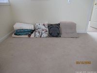Five folded fabric items on floor: a crocheted throw blanket in turquoise and beige, a cream-colored bedspread or blanket, and three decorative pillows of various designs including floral and medallion patterns.